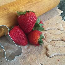 An image of Strawberry Yogurt Dog Treat dough, a bone cookie cutter, strawberries, and a rolling pin. The treats are made with diced strawberries, greek yogurt, whole wheat flour, ground flaxseed, steel cut oatmeal, and water.